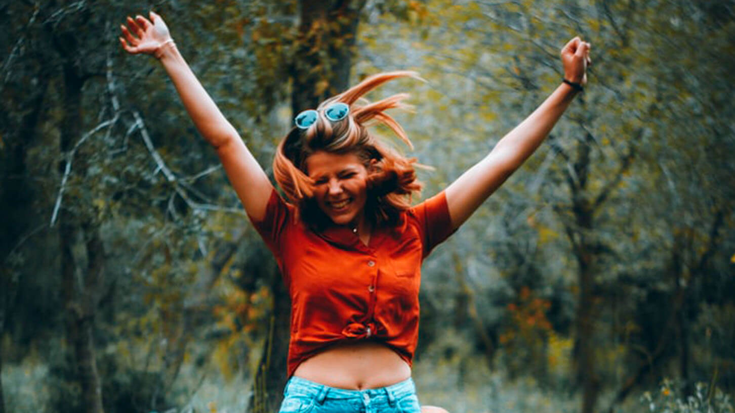 Ung kvinne hopper med hendene opp og med et stort smil, mens hun går i høstfarger ute i skogen.