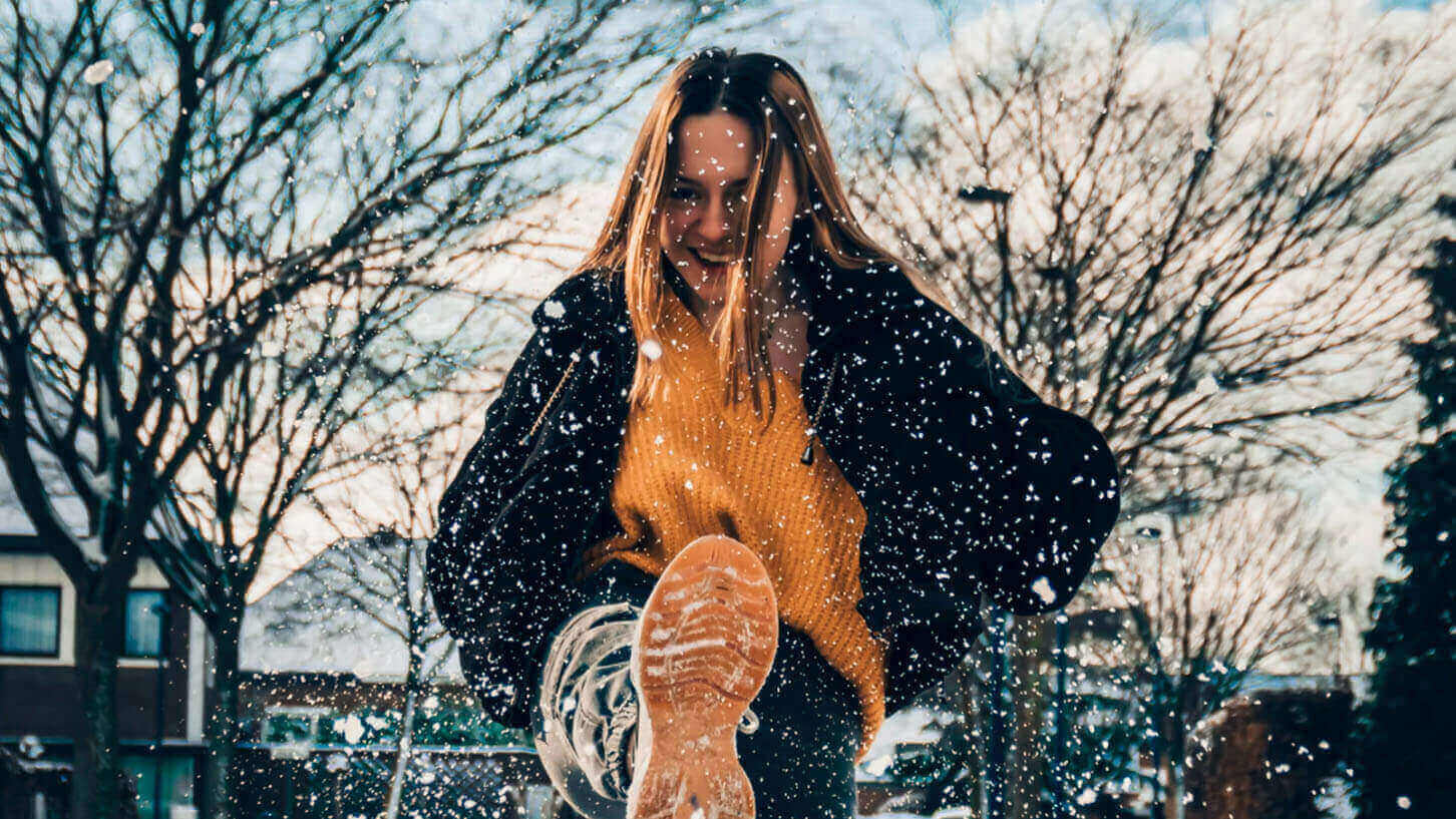 Ung dame i kledd vinterklær sparker snø mot kameraet med et smil om munnen.