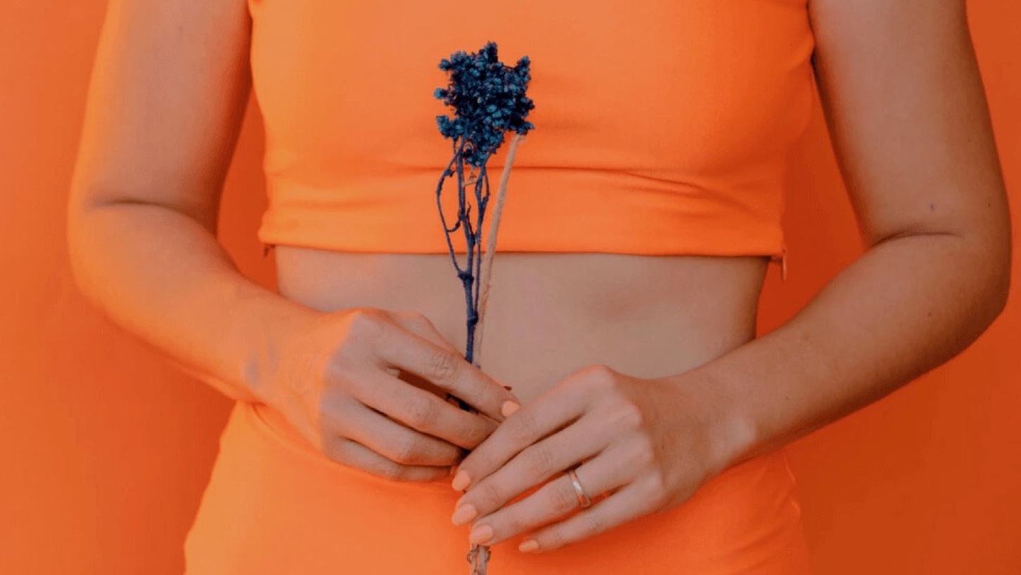 En dame i orange sportsklær holder en blå blomst foran en orange bakgrunn