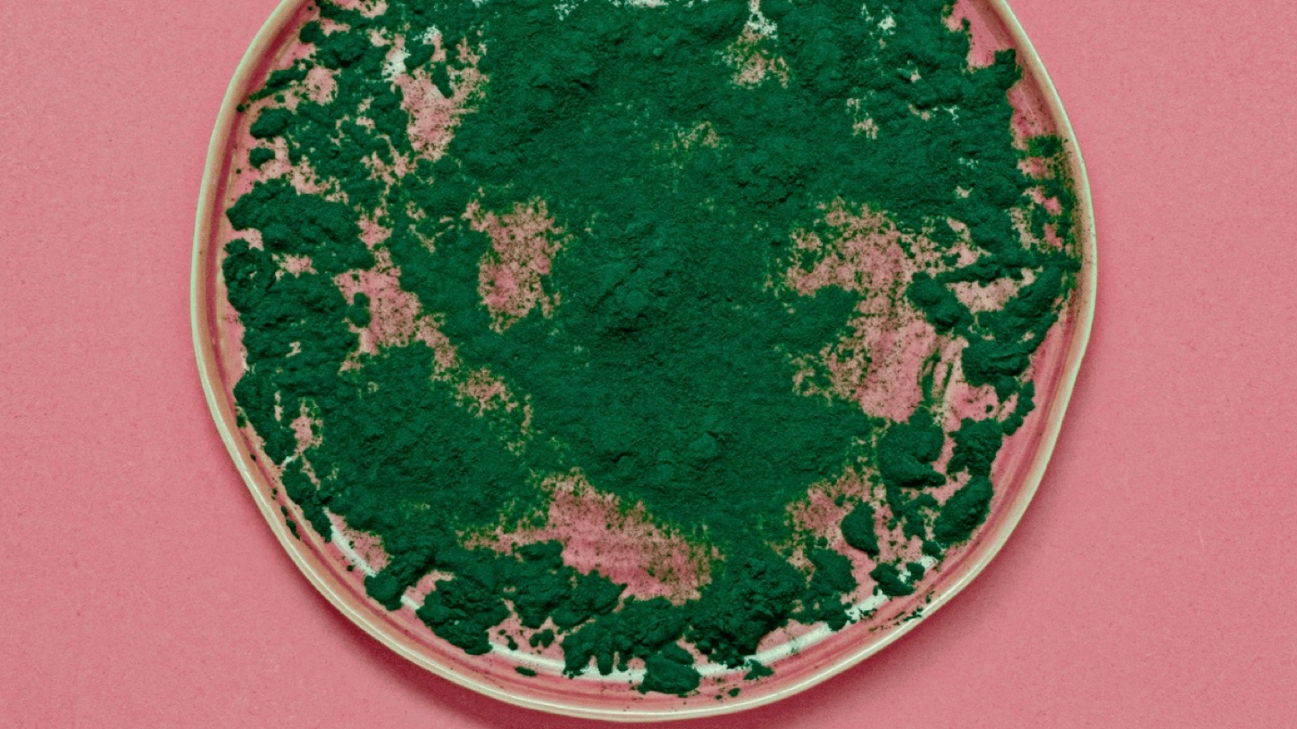 Grønn spirulina pulver på rosa tallerken som står på et rosa bord.