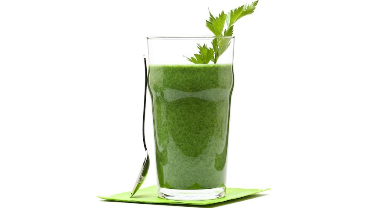 En grønn smoothie laget av supermat, i et høyt glass på en talerken, med en skje inntil glasset.