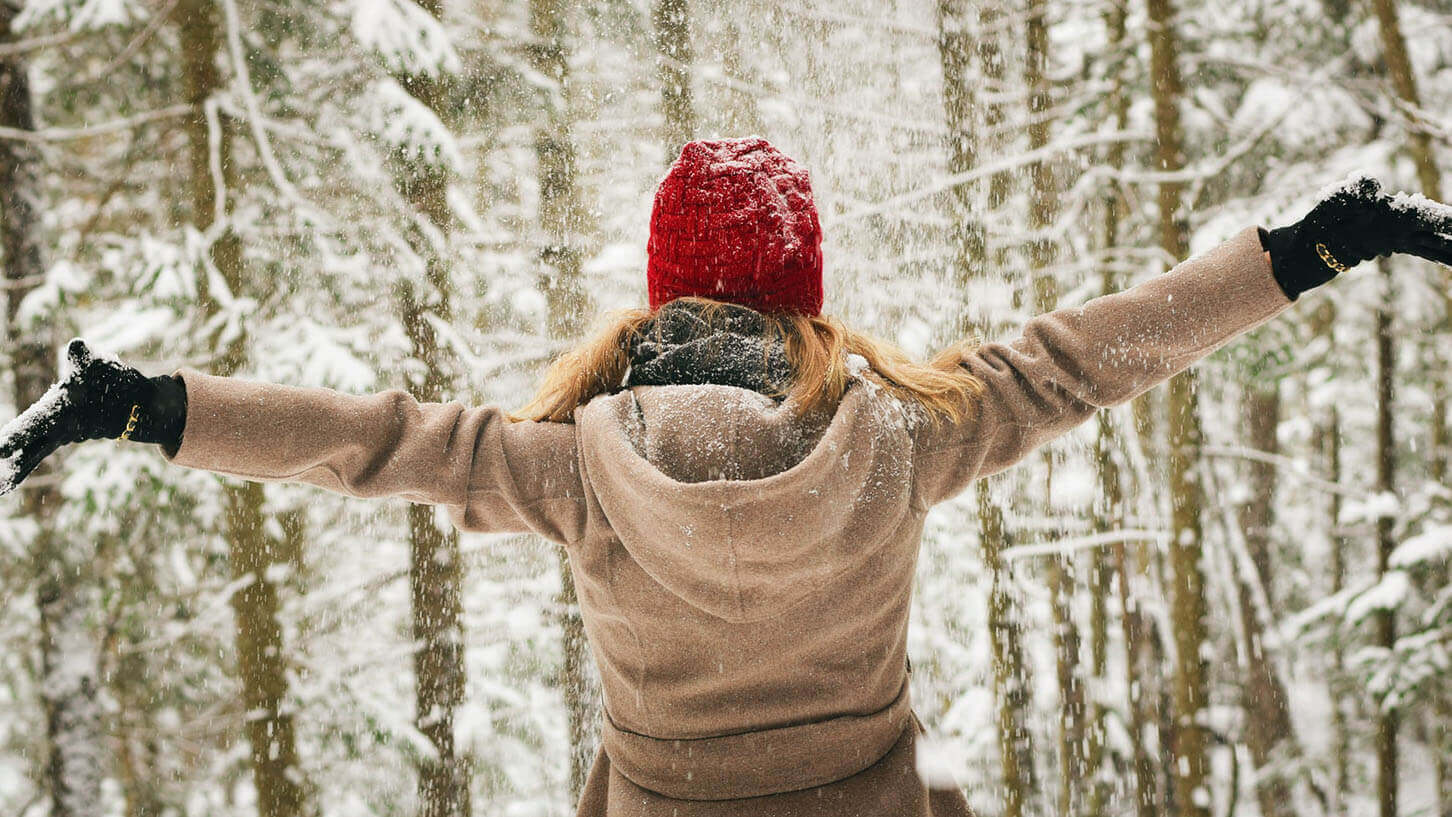 En kvinne med tykke vinterklær og rød lue står med ryggen til og armene ut, i skogen mens det snør.