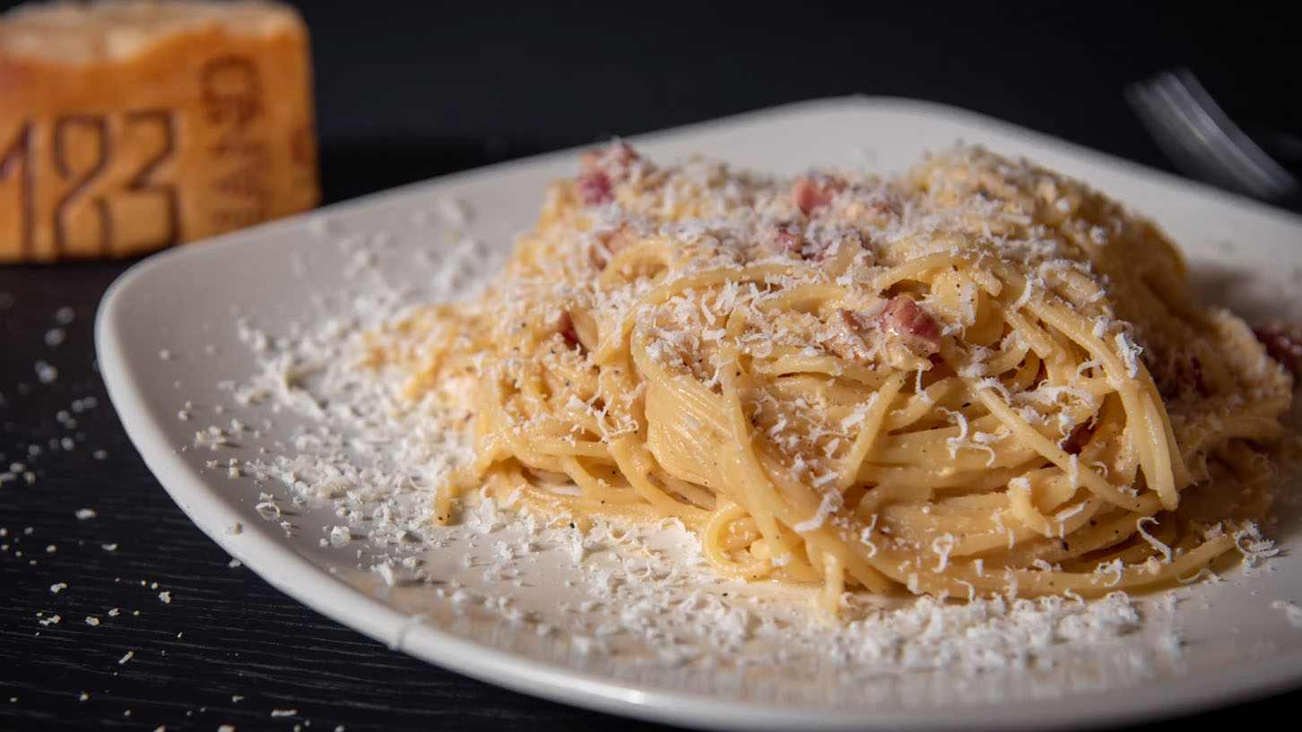 En hvit tallerken med saftig og fersk pasta carbonara med bacon toppet med masse parmasan, på et svart trebord.