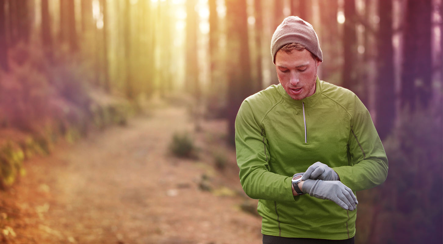 En ung mann sjekker pulsen på klokka mens han løper gjennom skogen i treningsklær